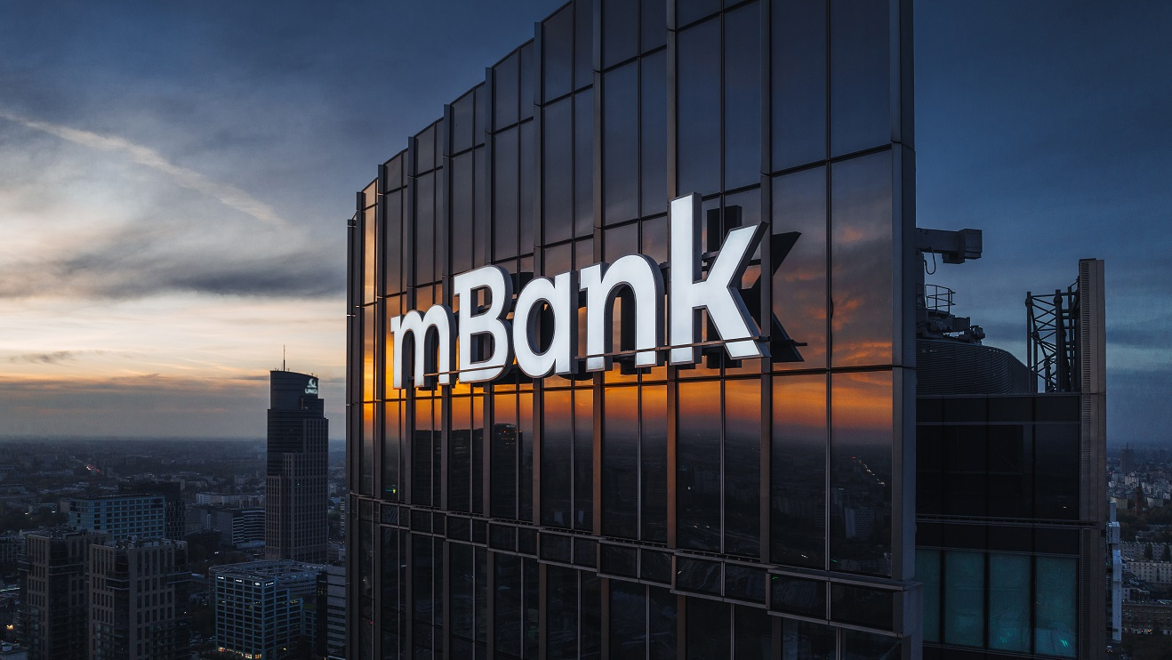 Widok na szklany budynek z podświetlonym logo mBanku o zmierzchu z widocznym zachodzącym słońcem i sylwetkami innych budynków w tle.