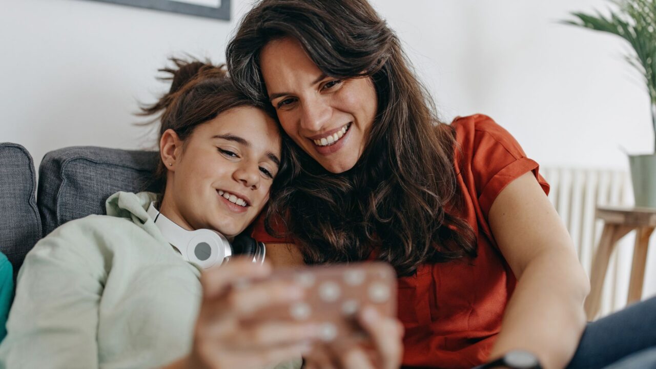 Eine Frau und ein junger Teenager lächeln, während sie auf einem Smartphone ein Selfie für die Instagram-Anwendung machen, während sie gemütlich auf einem Sofa in einem hellen Raum sitzen.