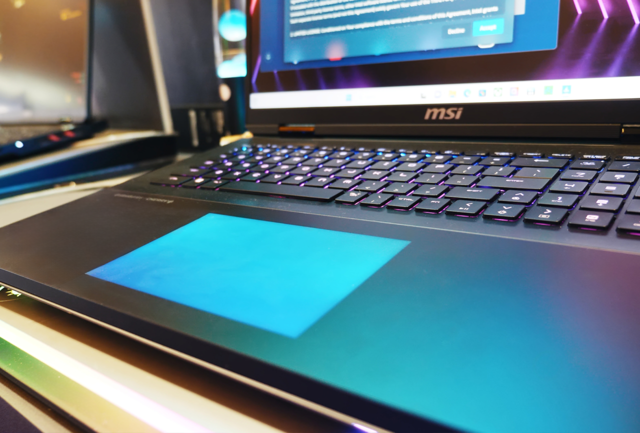 Laptop marki MSI z podświetlaną klawiaturą na pierwszym planie i włączonym ekranem w tle, na którym widać otwarte okno dialogowe.