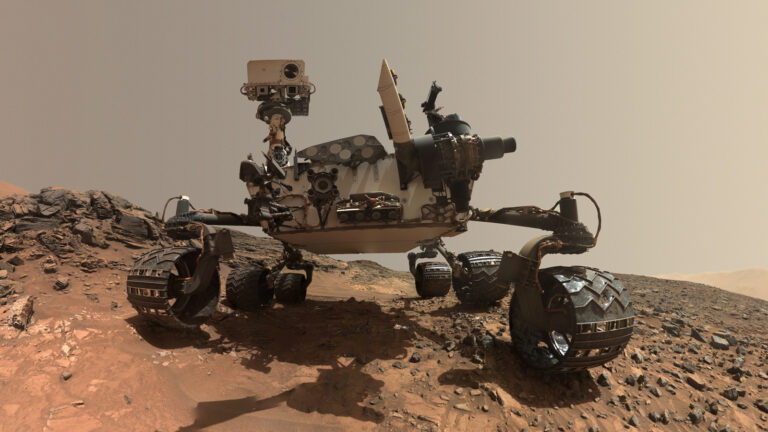 Łazik marsjański NASA Curiosity na powierzchni Marsa, z różnymi przyrządami naukowymi i kamerami, otoczony przez skaliste teren.