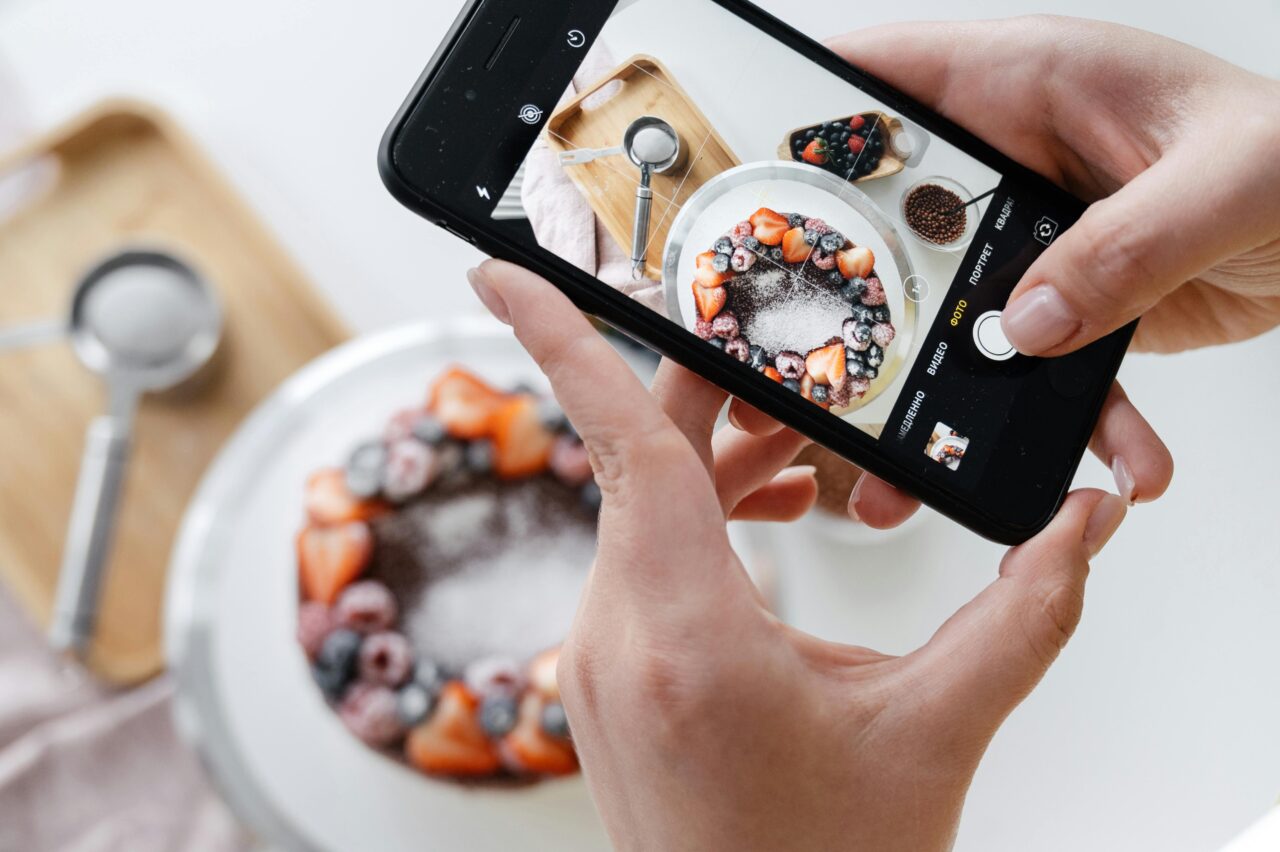 Osoba robi zdjęcie telefonem komórkowym zdrowego posiłku z jagodami i truskawkami posypanego cukrem pudrem, widok z góry na blat kuchenny.