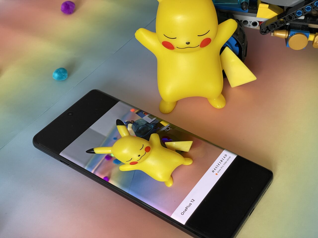 Zdjęcie figury Pikachu obok smartfona, na ekranie którego jest wyświetlone zdjęcie tej samej figurki Pikachu, z nieostrymi kolorowymi światłami w tle. Obok leży zabawka z klocków konstrukcyjnych.