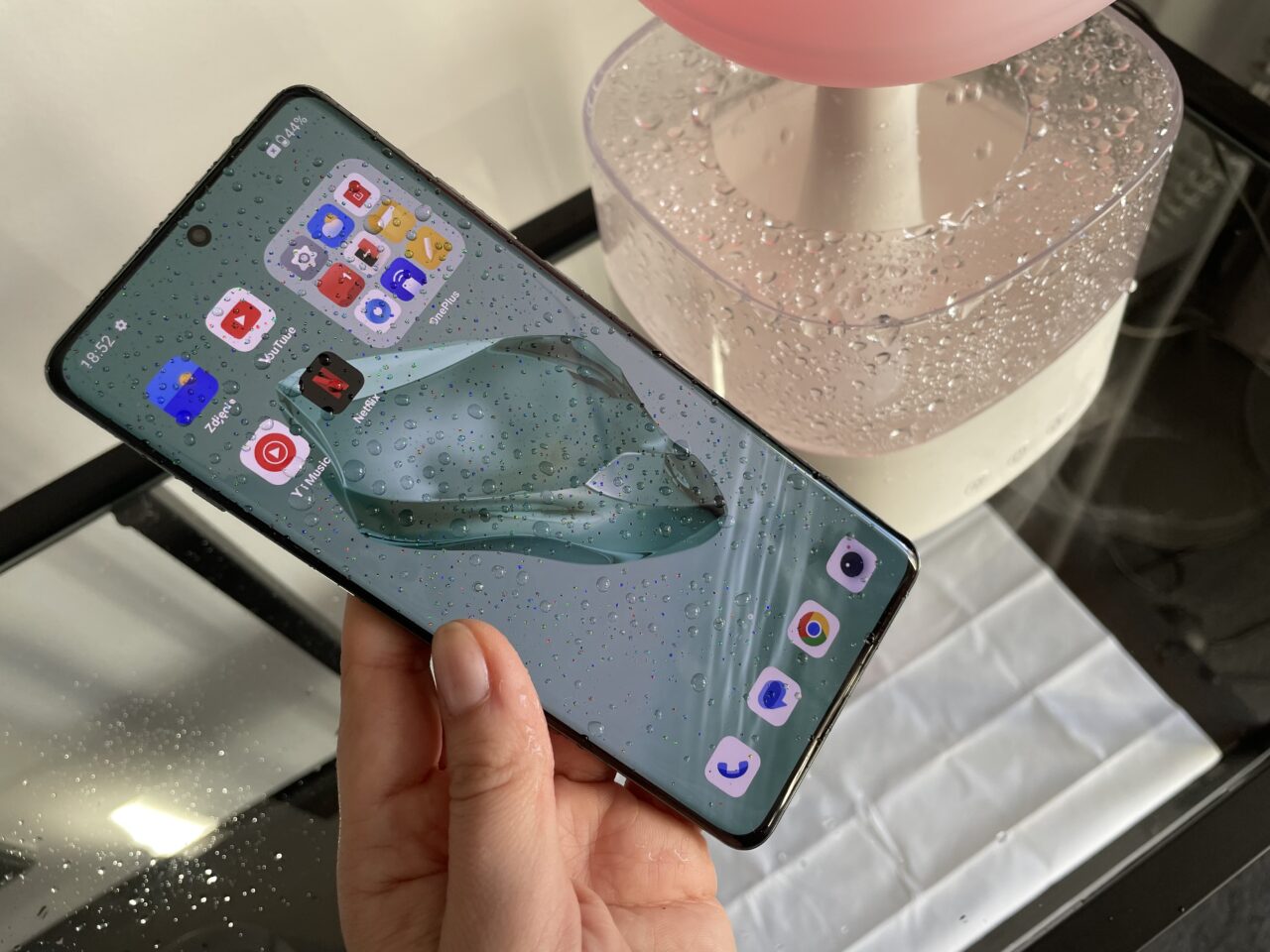 Osoba trzyma smartfon z aktywnym ekranem pokazującym ikony aplikacji na tle, które wygląda jak mokra powierzchnia z kroplami wody.