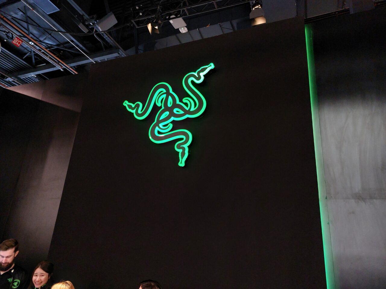 Zielone neonowe logo Razer na ciemnym tle, z ludźmi stojącymi poniżej.