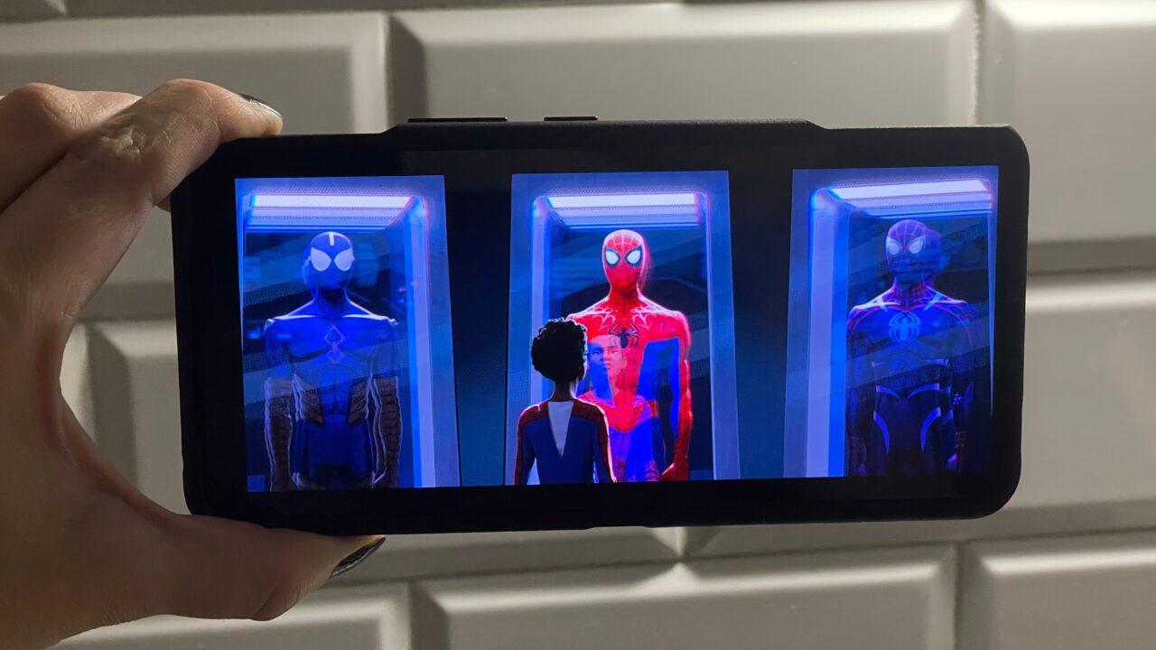 Osoba trzymająca telefon wyświetlający scenę z animowanego filmu przedstawiającego trzy postaci Spider-Mana w różnych kostiumach w komorze do testów.