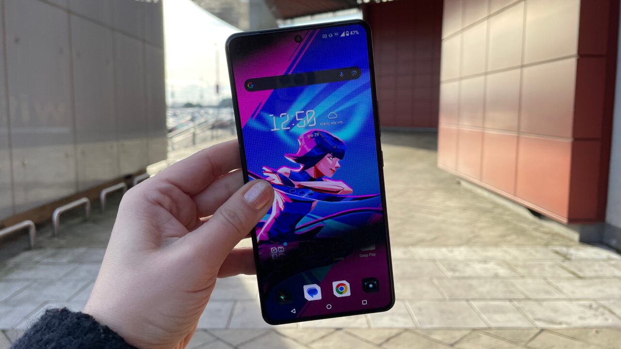 Dłoń trzymająca smartfon z ekranem wyświetlającym kolorową grafikę postaci z gry lub animacji w miejskiej scenerii.