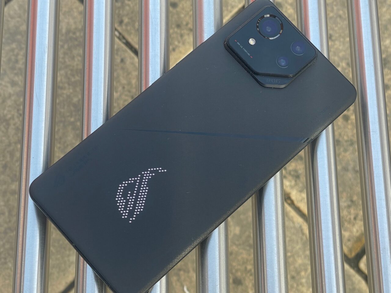 Czarny smartfon z logo w formie podświetlanego białego logotypu na tylnym panelu, położony na metalowych prętach.