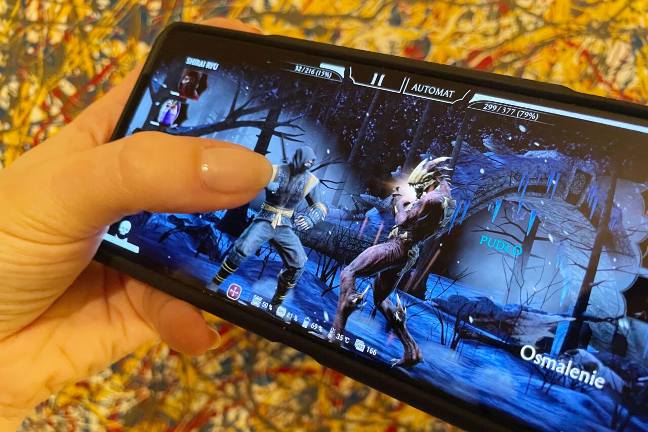 Osoba grająca na smartfonie w grę walki, gdzie dwie postacie starają się nawzajem pokonać w mrocznym lesie.