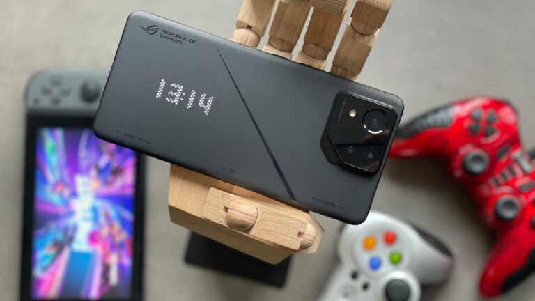 Czarny smartfon dla graczy z podświetlaniem "ROG" umieszczony na drewnianym stojaku, w tle rozmyta konsola Nintendo Switch i kontrolery do gier koloru czerwonego i białego.