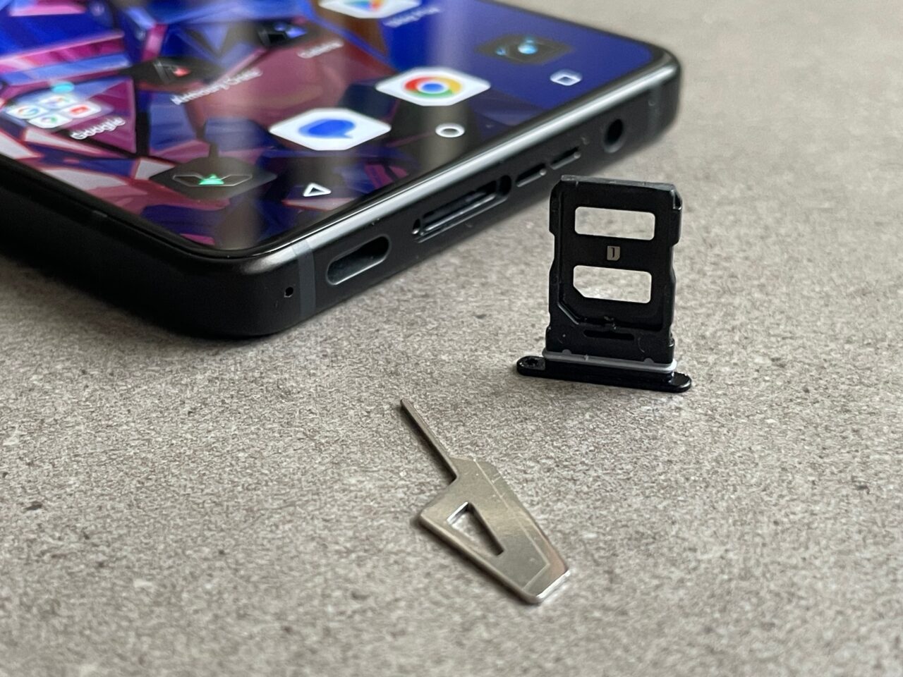 Częściowo widoczny smartfon leżący na szarym tle i obok niego leżąca tacka karty SIM oraz metalowe narzędzie do jej wyjęcia.