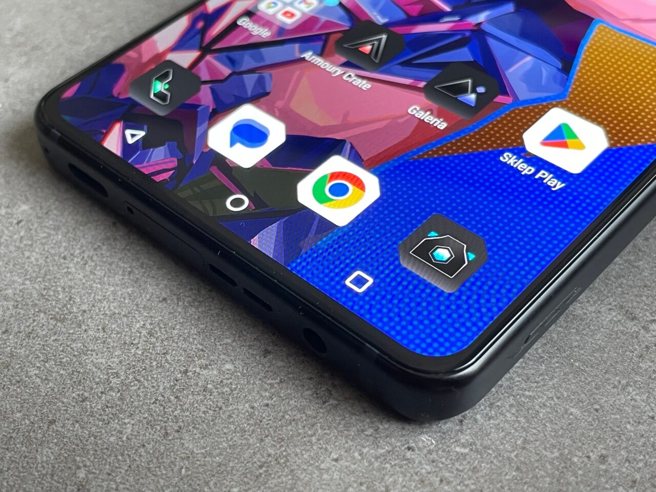 Część przednia smartfona z widocznymi kolorowymi ikonami aplikacji na tle abstrakcyjnej tapety, w tym ikona Sklepu Play, na blacie z szarą tkaniną.