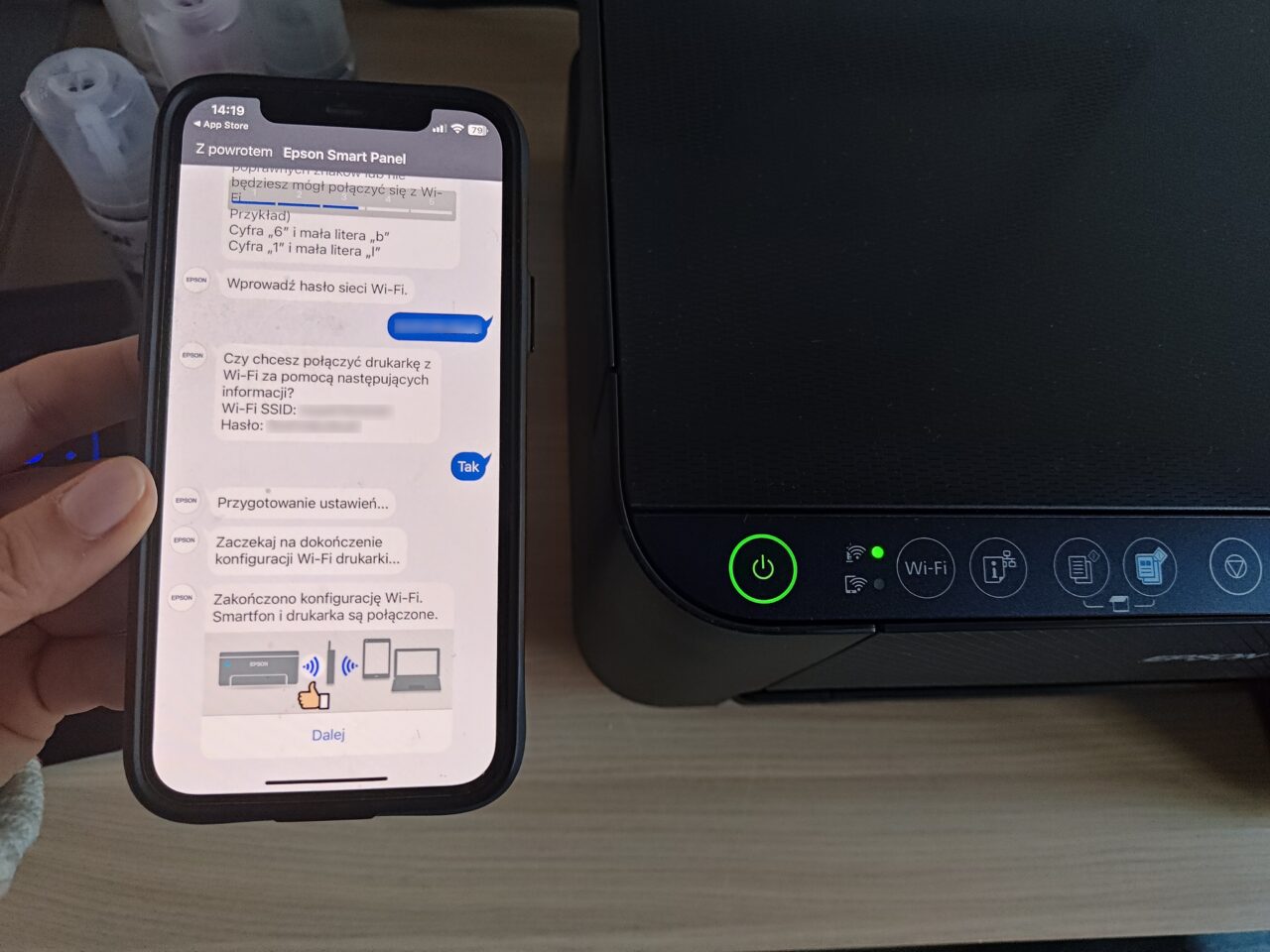 Dłoń trzymająca smartfon z otwartą aplikacją Epson Smart Panel obok drukarki z włączonym zielonym wskaźnikiem zasilania. Na ekranie smartfona proces konfiguracji Wi-Fi drukarki oraz wpisane hasło do sieci, częściowo zakryte niebieskim paskiem.