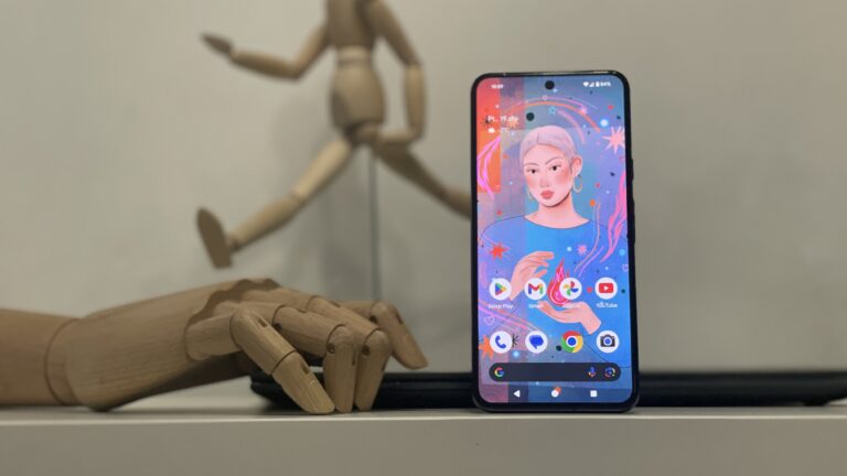 Smartfon z kolorową tapetą przedstawiającą grafikę kobiety, oparty o drewniane manekiny rąk, które znajdują się w rozmyciu w tle.