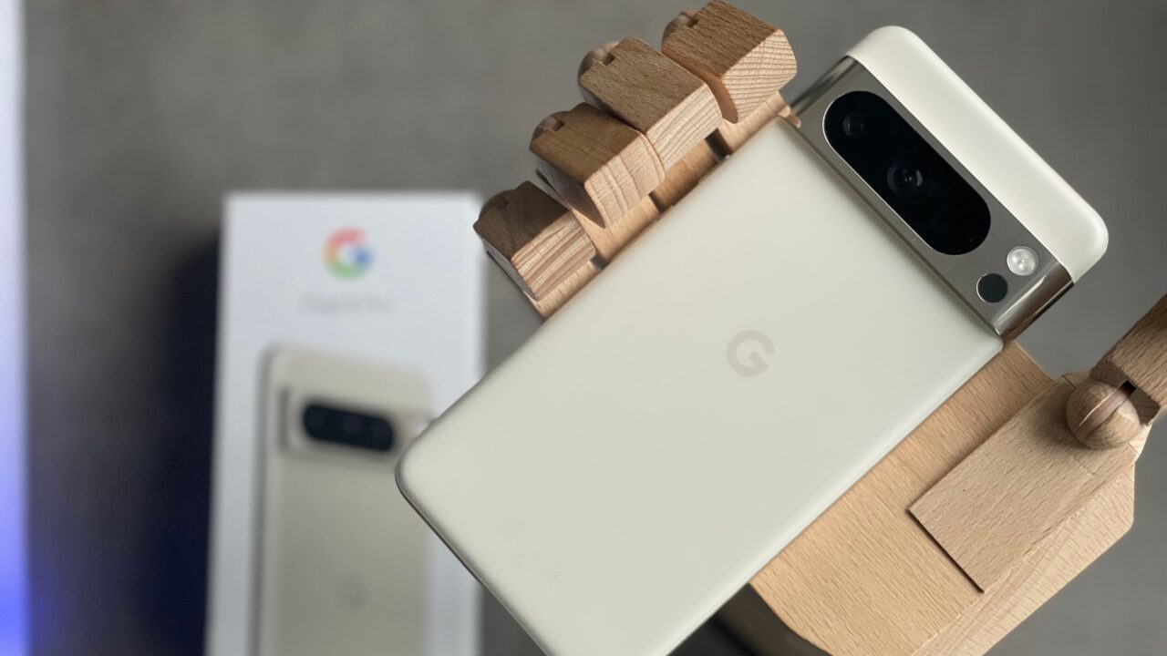 Smartfon Google Pixel 8 Pro w białym kolorze trzymany przez drewnianą dłoń manekina, oparty o oryginalne opakowanie.