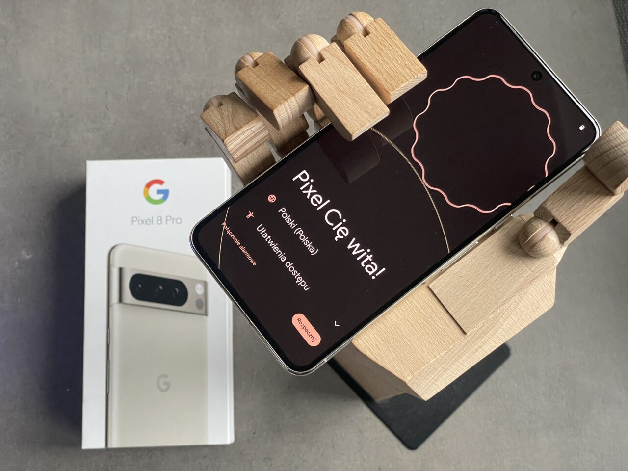 Smartfon Google Pixel 8 Pro na drewnianym stojaku, obok opakowanie produktu, na ekranie telefonu widoczny ekran konfiguracji WiFi.