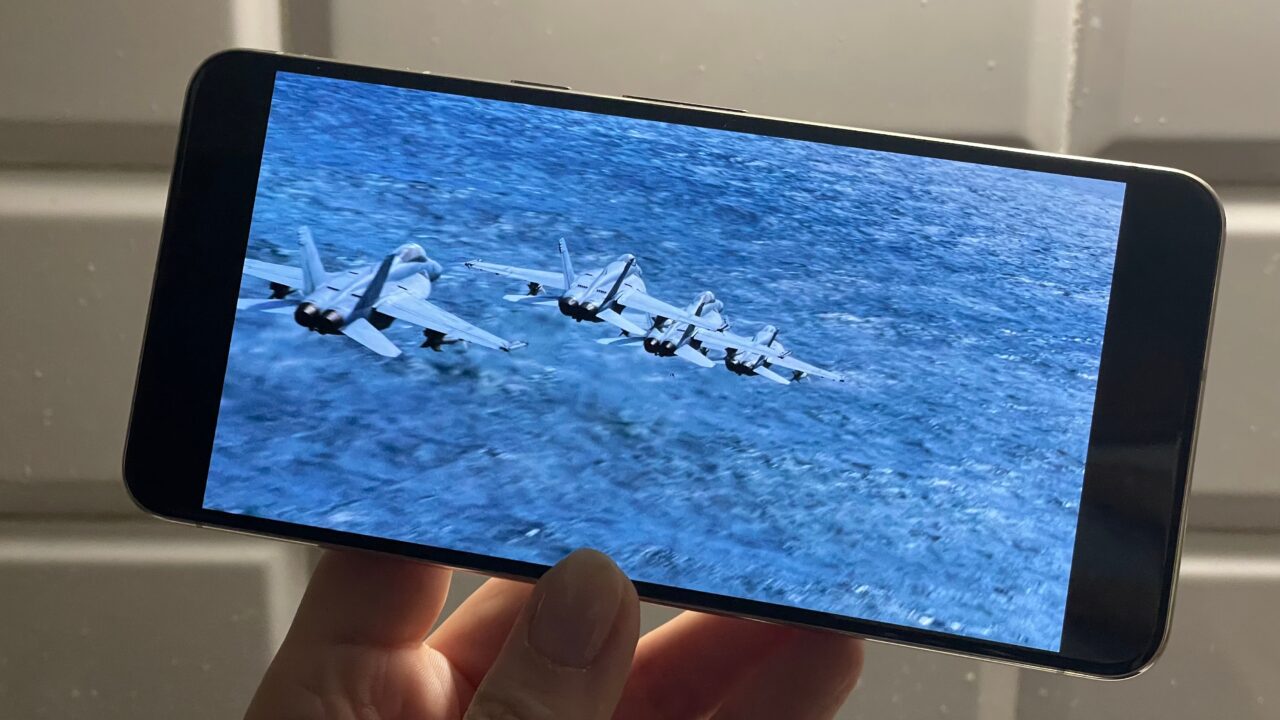 Osoba trzyma smartfon wyświetlający zdjęcie samolotów wojskowych lecących nisko nad powierzchnią wody.