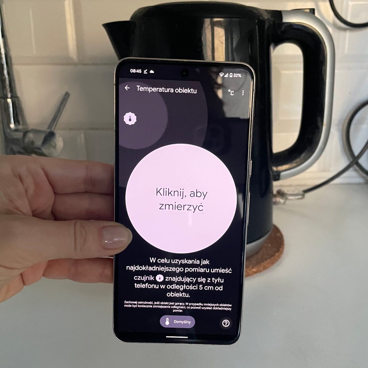 Osoba trzyma smartfon z otwartą aplikacją do mierzenia temperatury obiektów, z instrukcją "Kliknij, aby zmierzyć" na ekranie.