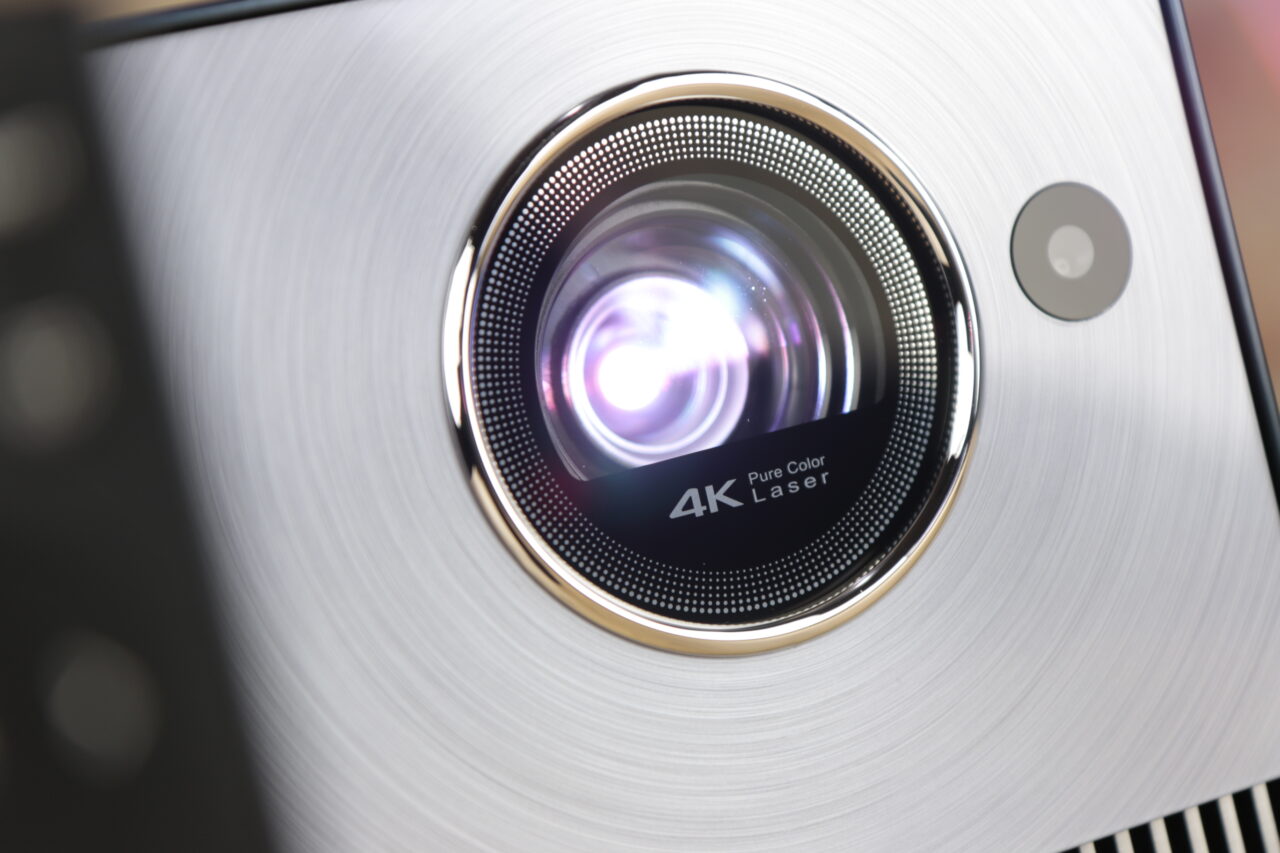 Obiektyw recenzowanego projektora Hisense C1 4K z napisem "Pure Color Laser" widoczny na metalicznym przedmiocie z okrągłym wzorem szczotkowanego aluminium.