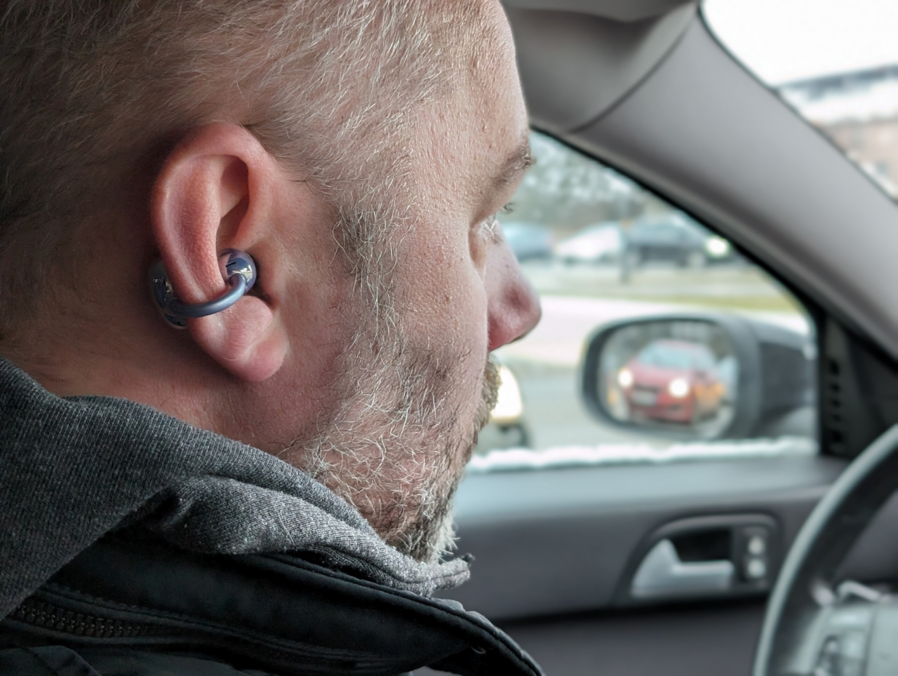 Mężczyzna z bezprzewodowym słuchawkiem w uchu siedzi w samochodzie, w tle widać lusterko boczne i inny samochód.