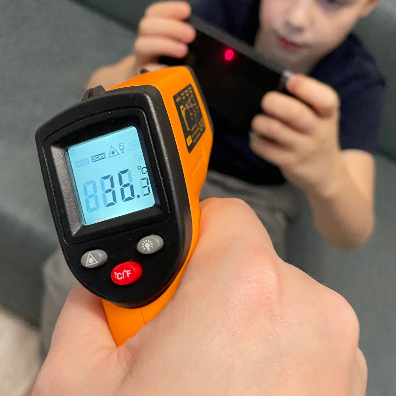 Dłoń trzymająca termometr podczerwieni pokazujący temperaturę 83,3°C, w tle rozmyty wizerunek dziecka patrzącego na smartfon.