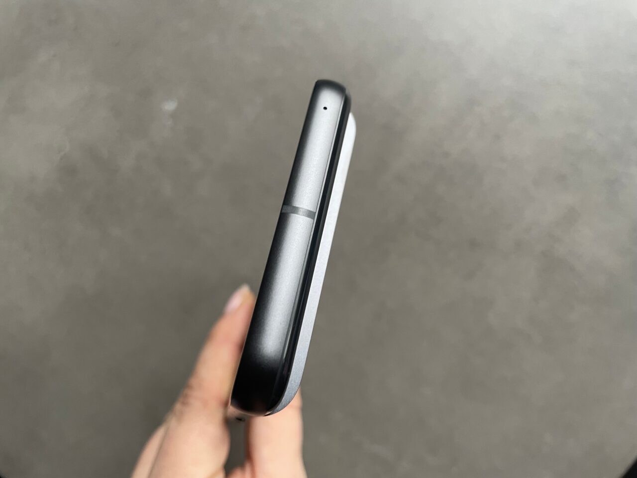 Czarny smartfon trzymany w dłoni na szarym tle, widok boczny ukazujący krawędź i część ekranu.