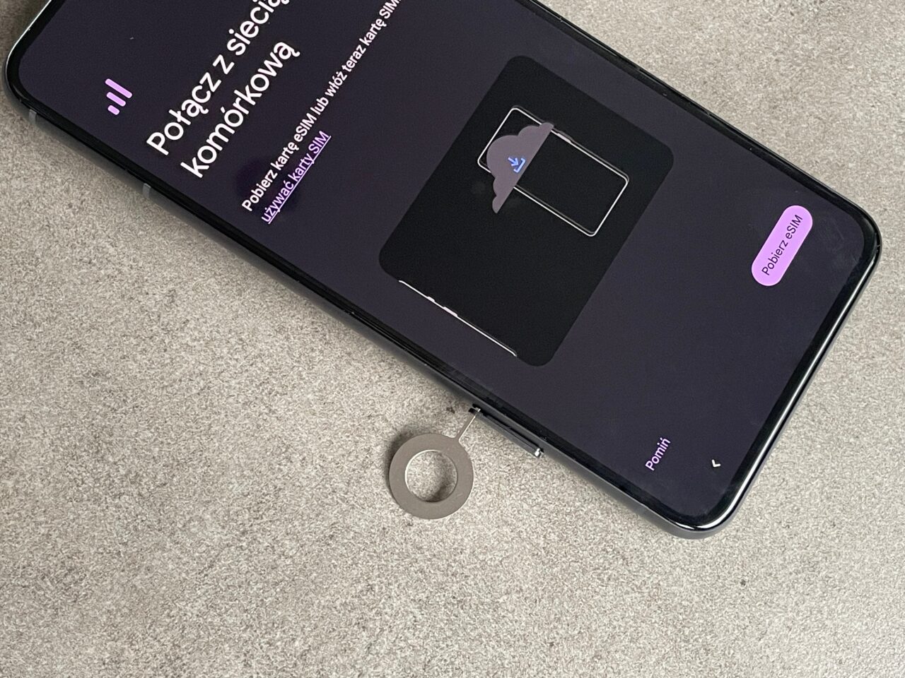 Czarny smartfon leży na szarym podłożu z ekranem prezentującym wiadomość o braku karty SIM i grafiką karty SIM w środku, obok dwie naklejki z napisem "Podłącz się" i "Pomiń".
