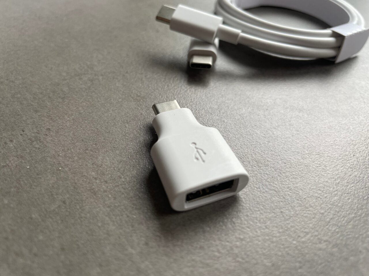Biały adapter USB-C na szarym tle z widocznym logo USB i zgietym białym kablem z wtyczką USB-C w tle.