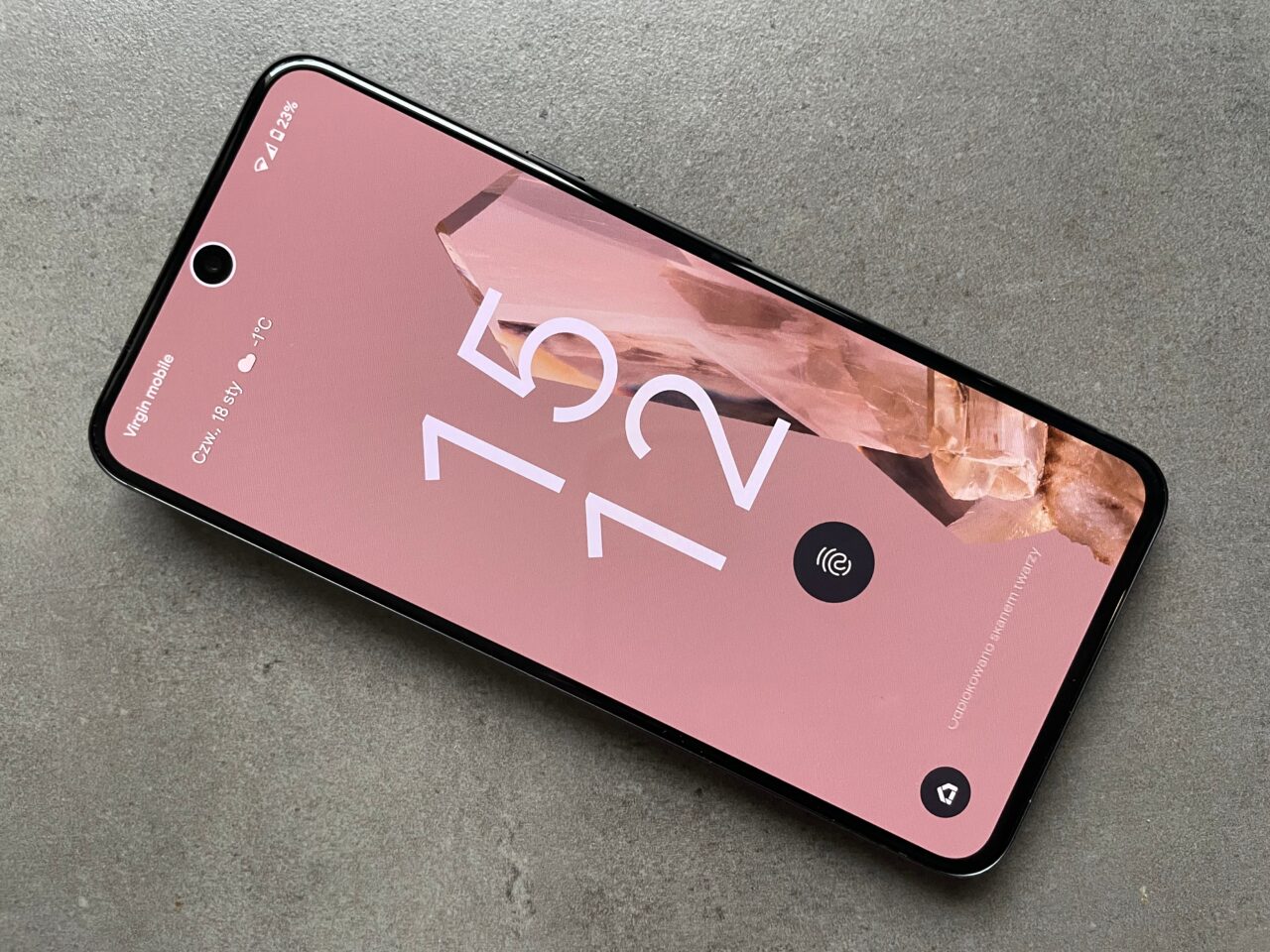 Smartfon w kolorze różowym leżący na szarym tle, z widocznym ekranem blokady i dużymi cyframi 12:45 na tle różowych kryształów.