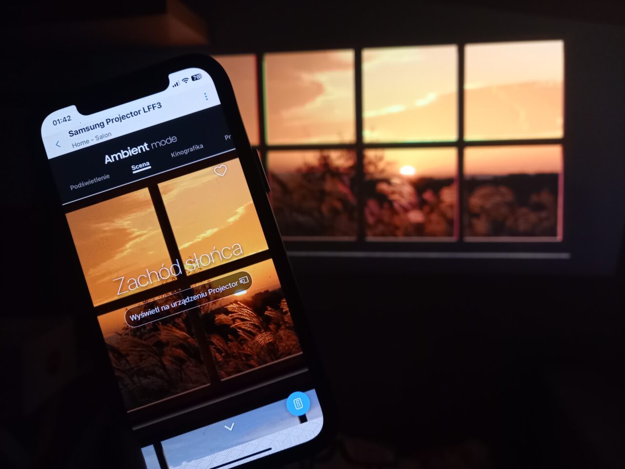 Smartfon wyświetlający obraz zachodu słońca z napisem "Zachód słońca", trzymany przed projekcyjnym ekranem z tym samym obrazem w tle.
