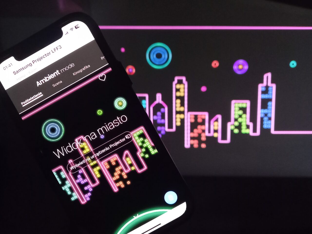 Ekran smartfona wyświetlający opcje projektora z napisem "Ambient mode" i tłem w postaci kolorowej grafiki miasta wyświetlanej przez projektor.