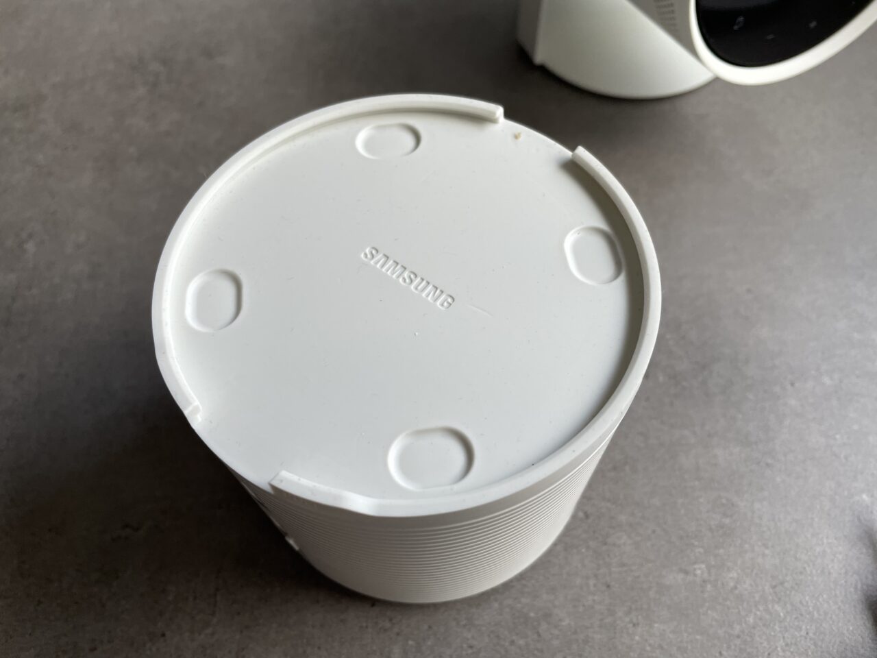 Biała plastikowa pokrywka z logo firmy Samsung na szarym tle obok fragmentu urządzenia w kolorze białym.