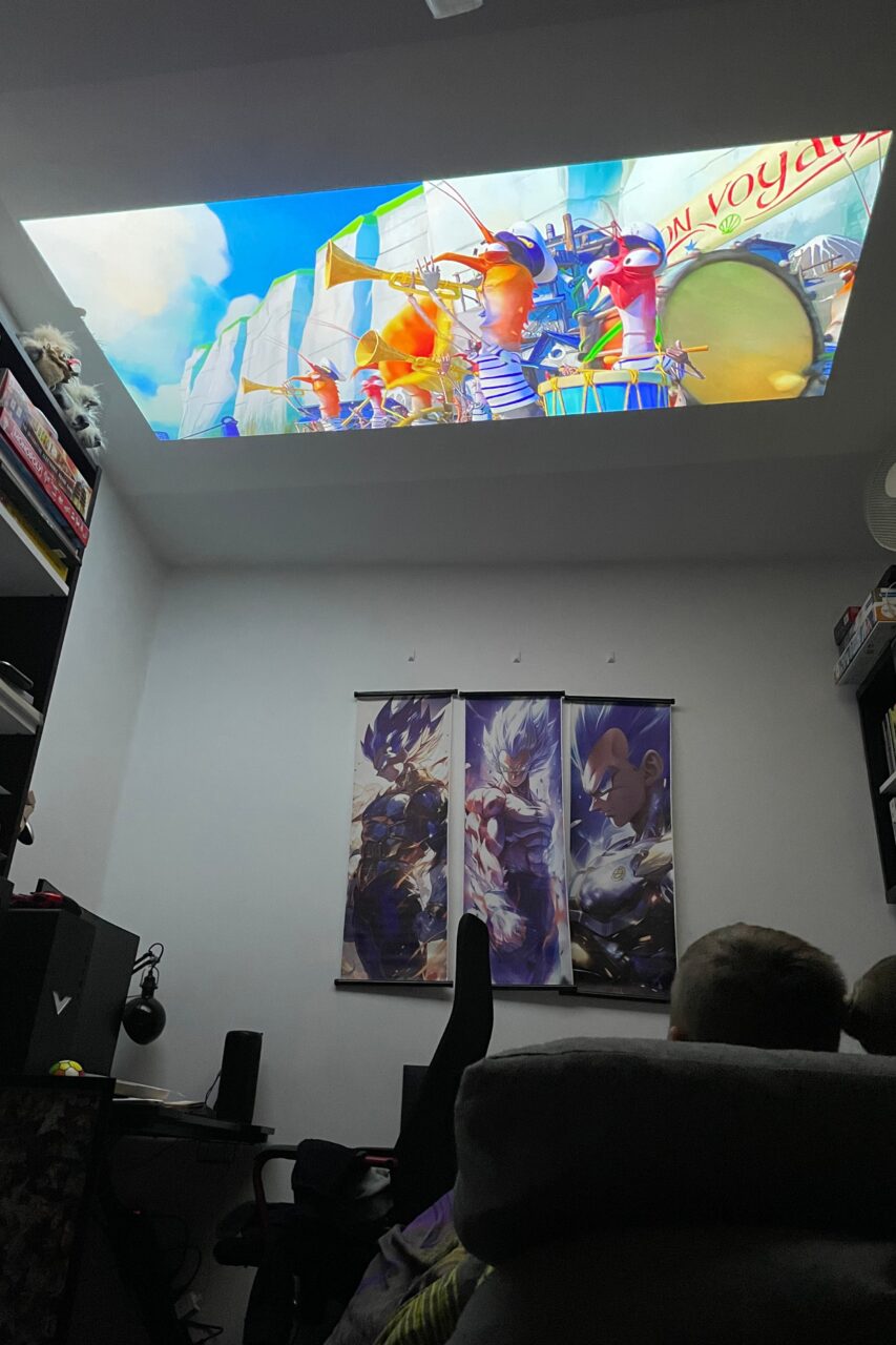 Pokój z projektorem wyświetlającym animację na ścianie, podświetlane plakaty z postaciami z anime na ścianie oraz widoczna tylna część głowy osoby siedzącej na fotelu.