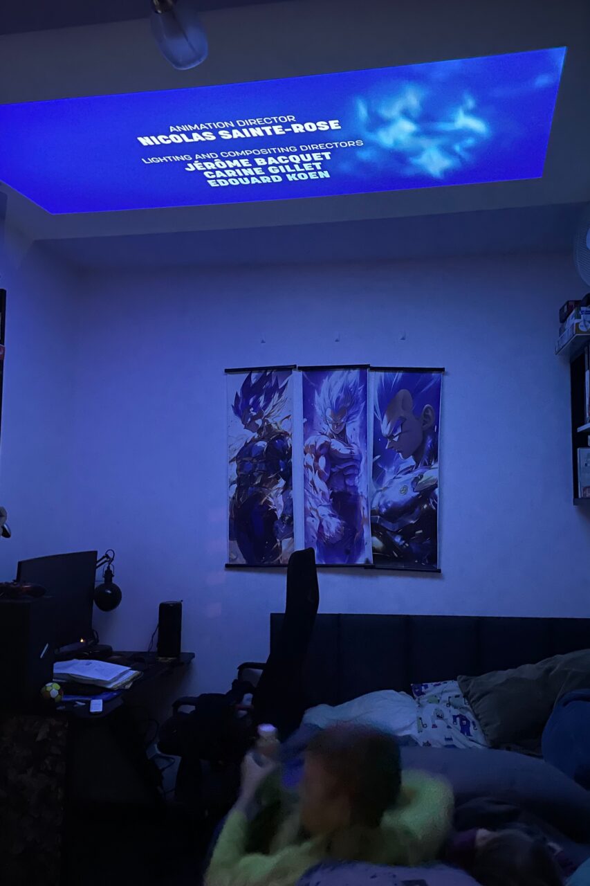 Pokój z niebieskim oświetleniem LED, projekcją napisów końcowych na suficie, dwuczęściowym obrazem z postaciami anime na ścianie, fotelami komputerowymi i osobą leżącą na kanapie.
