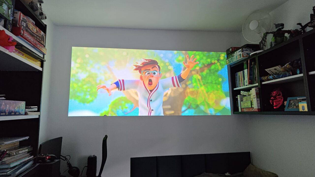 Pokój z projektorem wyświetlającym animowaną postać wyrażającą zdziwienie na ekranie, otoczony półkami z książkami i grami planszowymi.