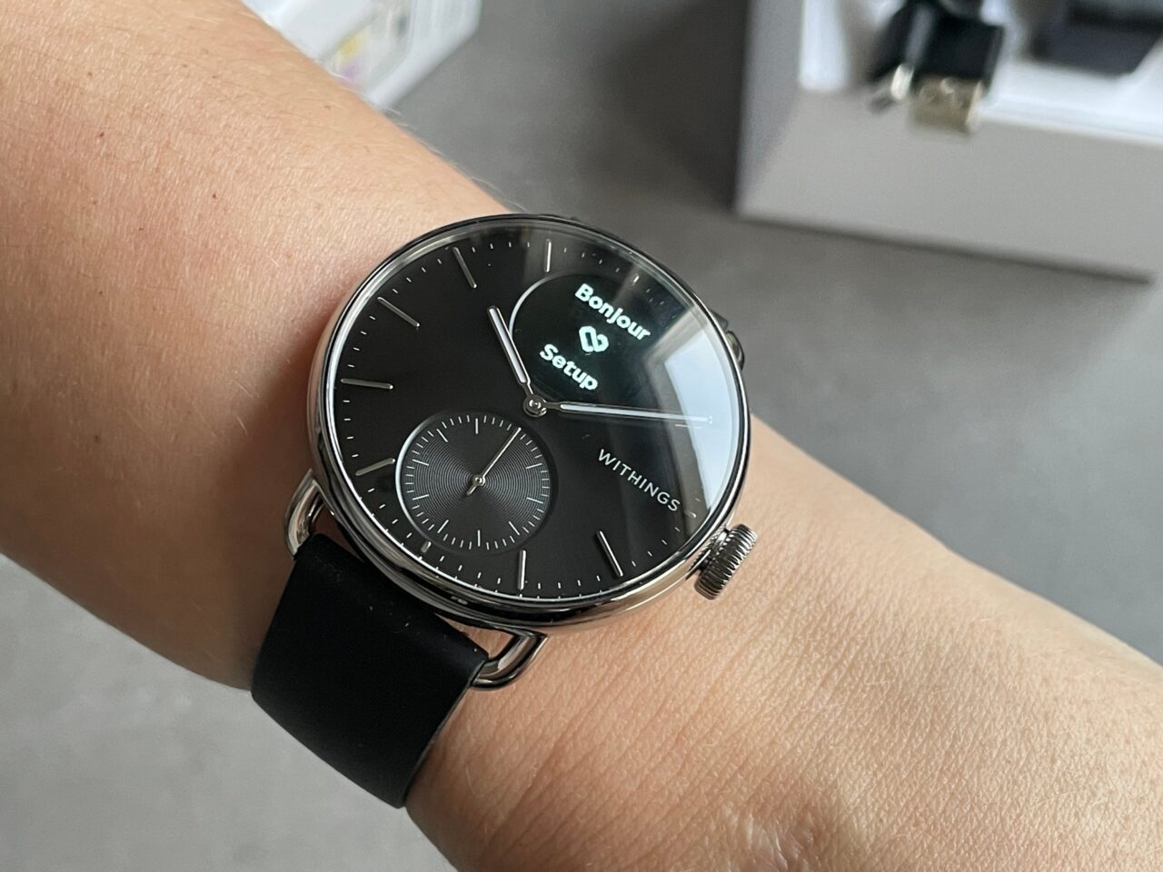 Smartwatch z czarnym paskiem na nadgarstku osoby, wyświetlający czas i powiadomienie z napisem "Bonjour".