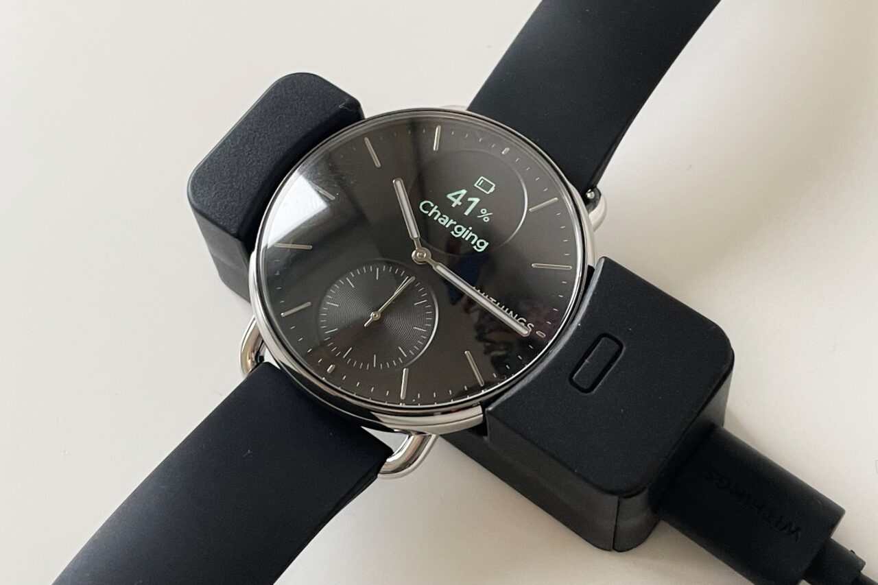 Czarny smartwatch z analogowym wyglądem tarczy naładowany na czarnej ładowarce, wyświetlający stan baterii na poziomie 41% z napisem "Charging".