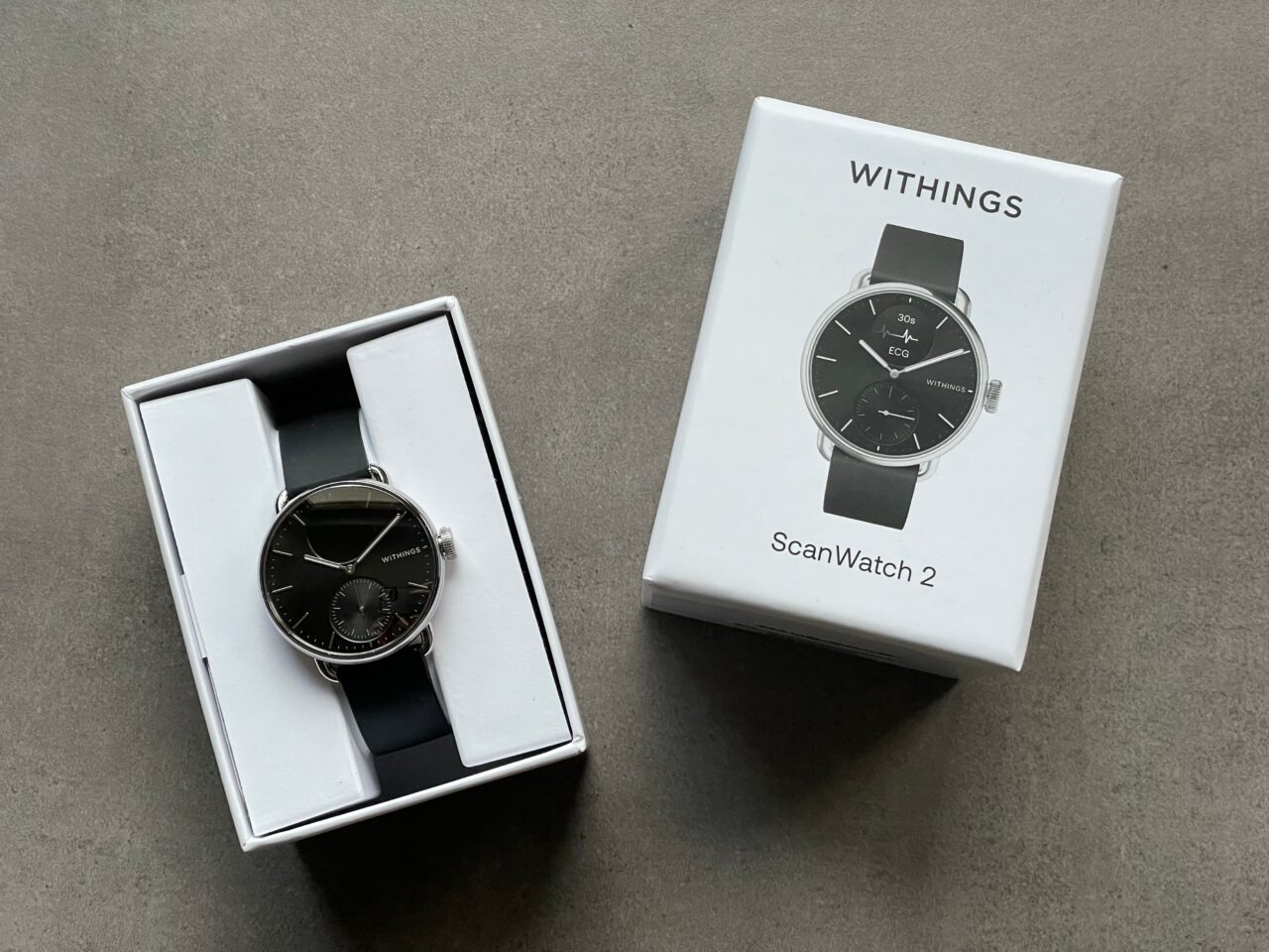 Zegarek Withings ScanWatch 2 leżący w otwartym pudełku obok zamkniętego opakowania, na szarym tle.