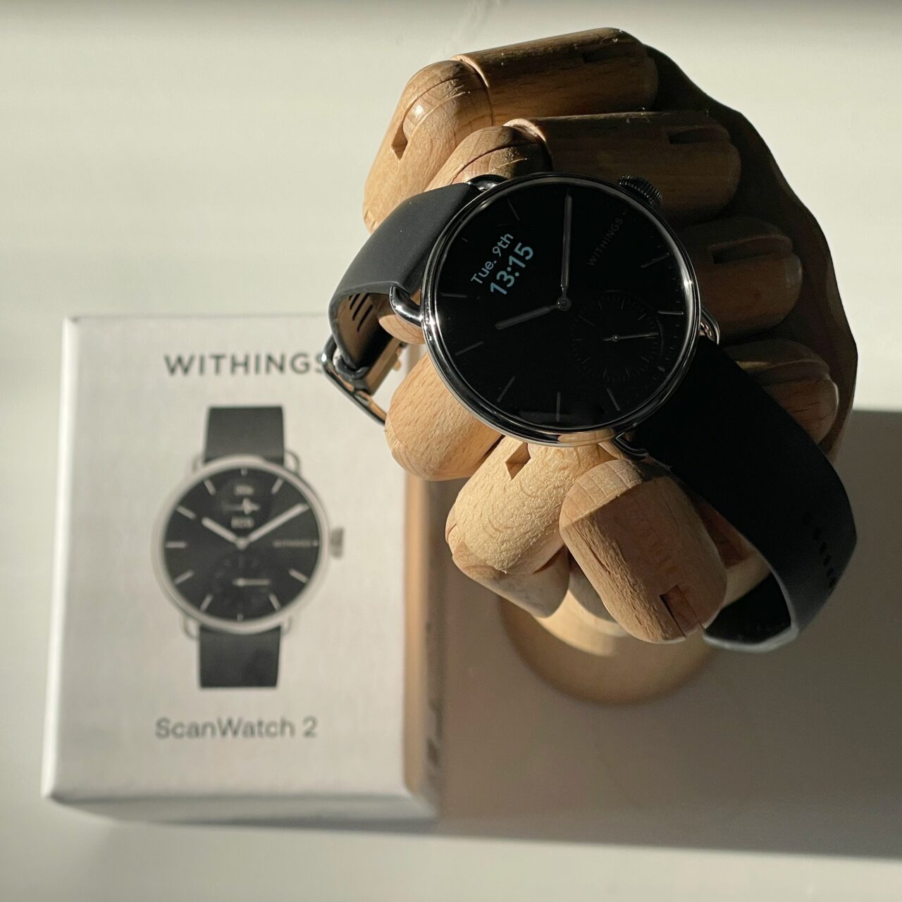 Czarny zegarek Withings ScanWatch na drewnianej dłoni manekina, z pudełkiem produktu w tle.