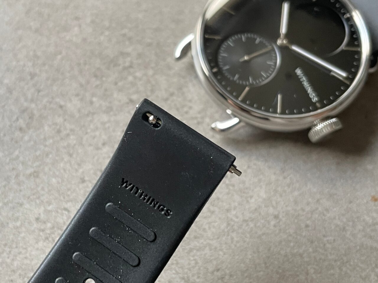 Czarny pasek zegarka z logo "WITHINGS" w dolnej części zdjęcia i częściowo widoczny srebrny zegarek z czarną tarczą w tle.
