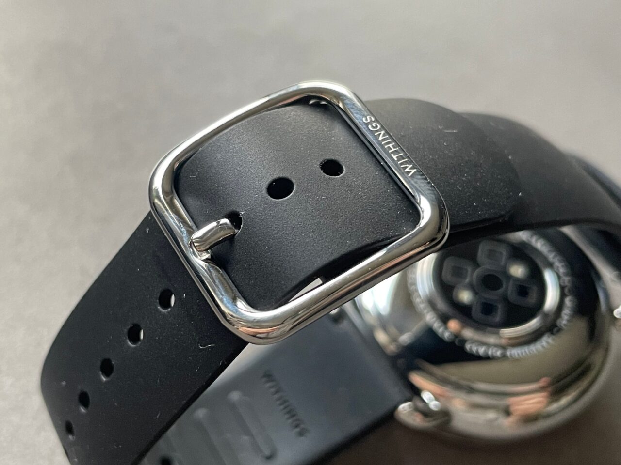 Czarny gumowy pasek do smartwatcha z metalową klamrą, w tle widoczna odwrotna strona innego smartwatcha.