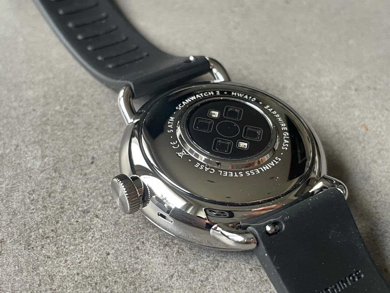 Zegarek inteligentny odwrócony do góry dnem na tle o szarej fakturze, pokazujący spód urządzenia z sensorami i informacjami o modelu.