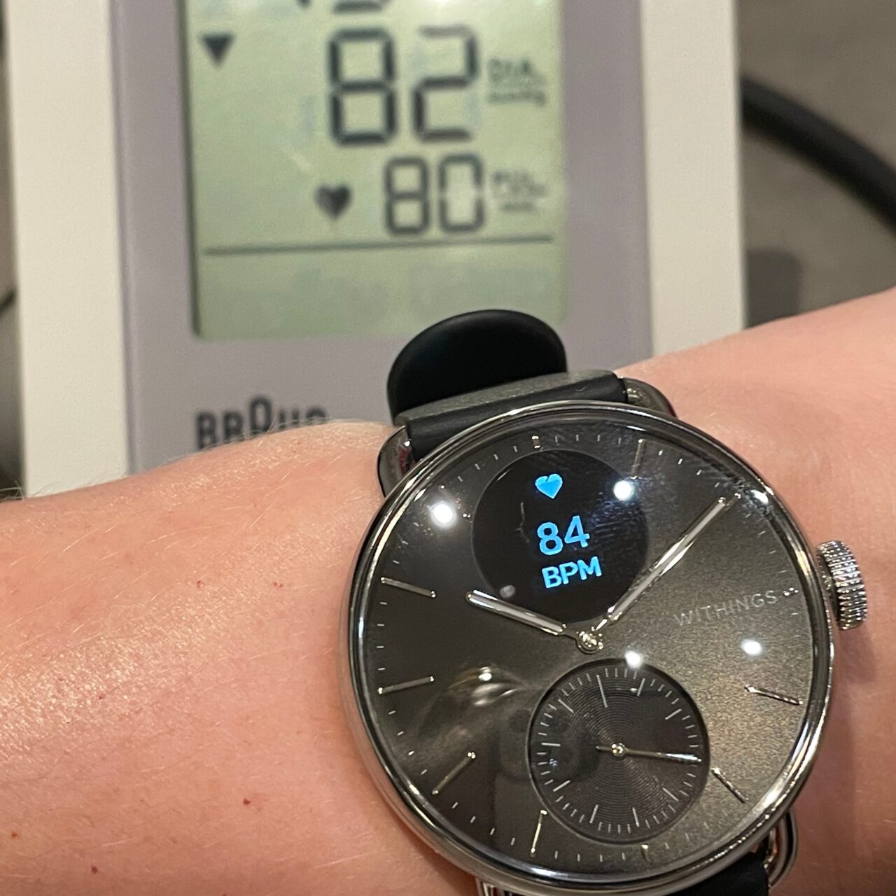 Zdjęcie nadgarstka osoby z inteligentnym zegarkiem Withings pokazującym tętno 84 BPM na tle monitora ciśnienia krwi wyświetlającego również tętno 82 BPM.