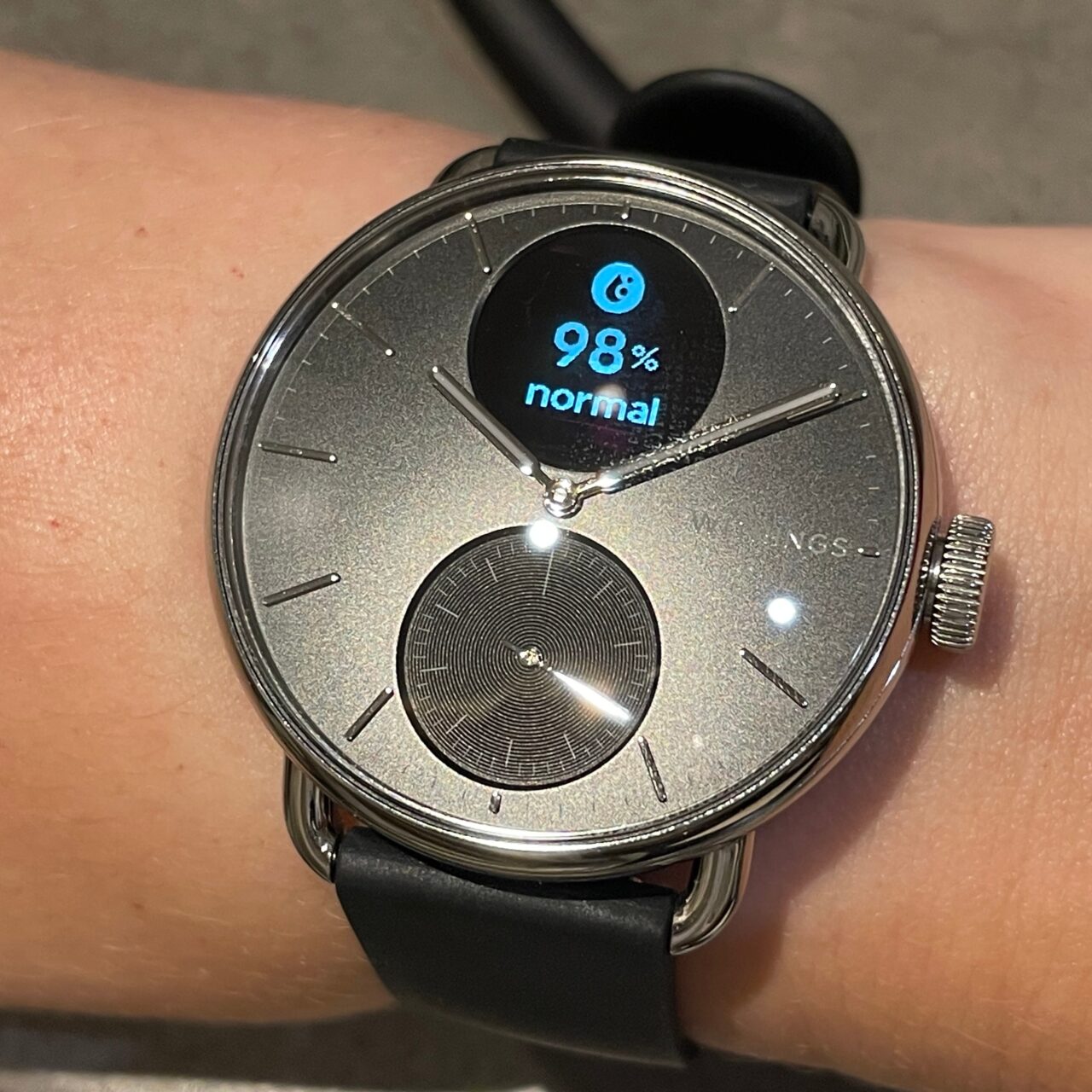 Zegarek na ręku z cyfrowym wyświetlaczem pokazującym 98% i słowo "normal" na tarczy analogowej.