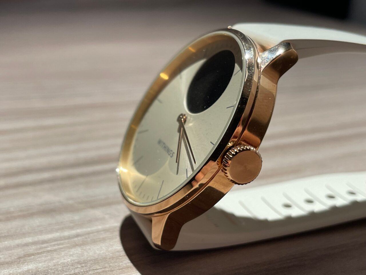 Zegarek na białym pasku położony na drewnianym blacie, złota tarcza odbija światło, widoczna koronka do ustawiania czasu.