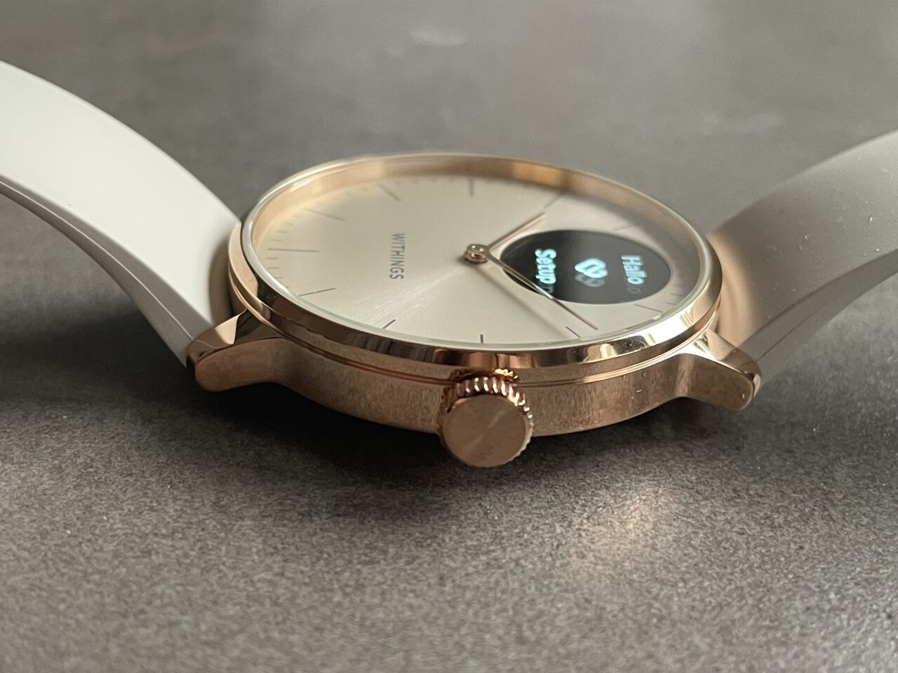 Zegarek na szarym tle z częściowo widoczną tarczą cyfrową i logo na analogowej tarczy, złotą kopertą i białym paskiem.