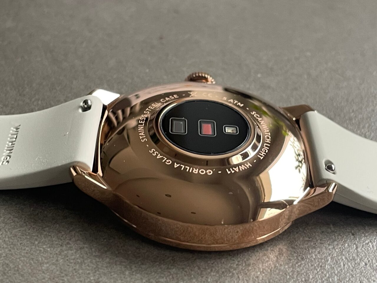 Różowe złoto smartwatch z wyświetlaczem odwróconym do dołu, pokazujący tylną część z czujnikami i napisami, na szarym tle.