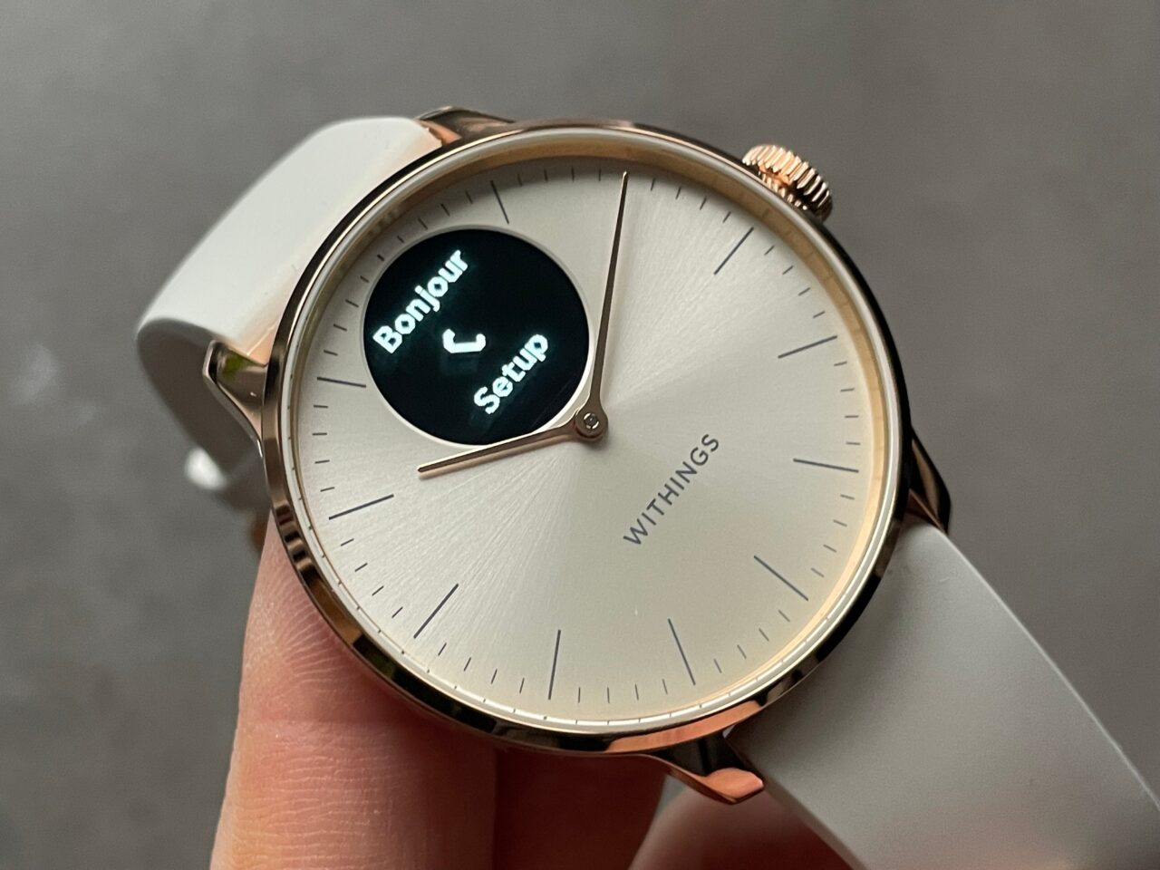 Zegarek z analogową tarczą w kolorze jasnoszarym i różowo-złotą obudową, wyświetlający cyfrowo napis "Bonjour Setup" na małym okienku OLED, z logo "WITHINGS" na dole tarczy.
