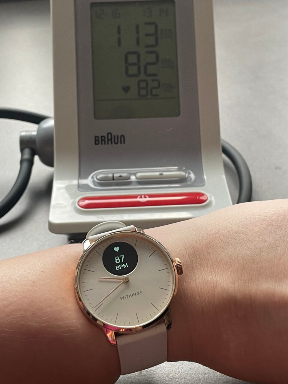 Nadgarstek z założonym smartwatchem Withings pokazującym tętno 87 BPM na pierwszym planie i ciśnieniomierz Braun w tle wyświetlający ciśnienie krwi 113 na 82 oraz tętno 82.