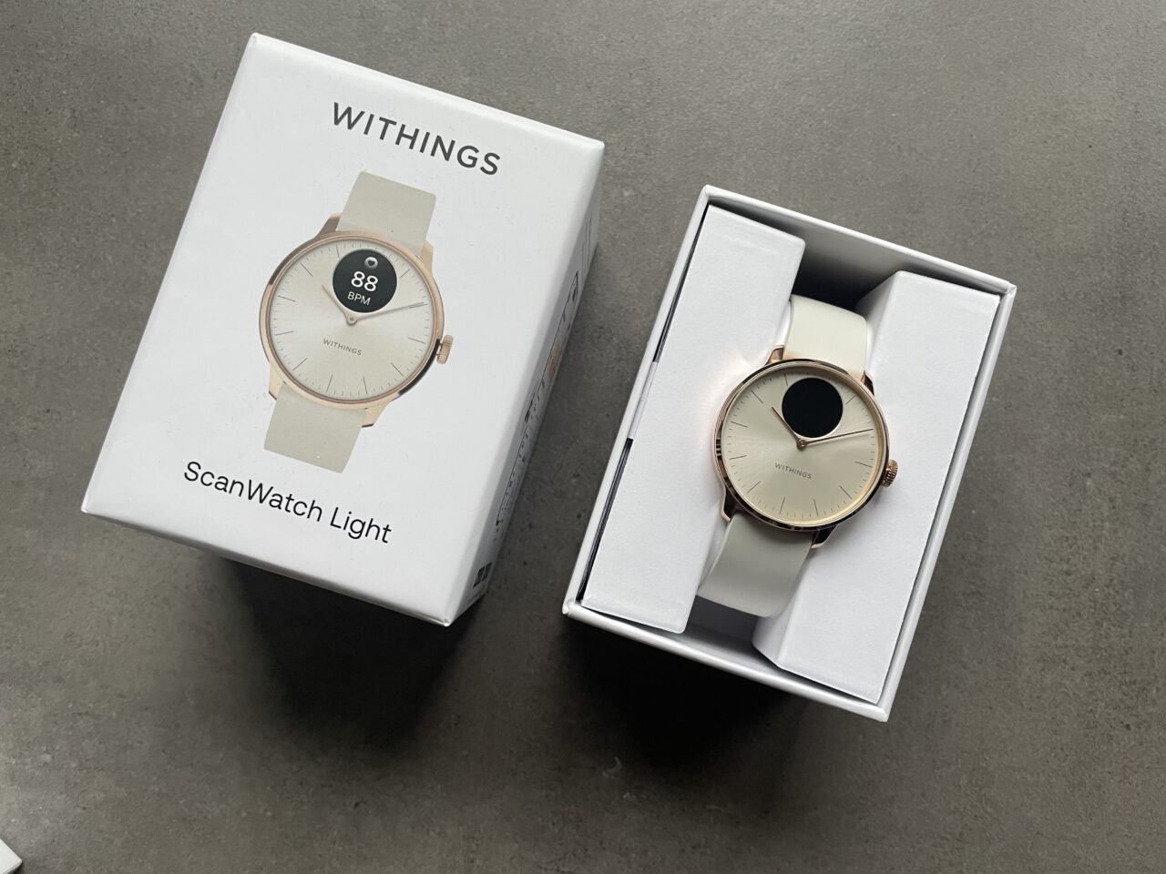 Zegarek Withings ScanWatch Light w różowo-złotej obudowie na białym pasku, umieszczony w otwartym białym pudełku, obok którego leży zamknięte pudełko z nadrukowanym obrazem tego zegarka.
