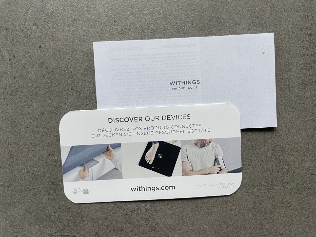 Broszura produktowa Withings leżąca na szarej powierzchni z napisem "DISCOVER OUR DEVICES" w trzech językach oraz zdjęciami osób używających urządzenia medyczne, a nad nią umieszczony przewodnik produktowy Withings.